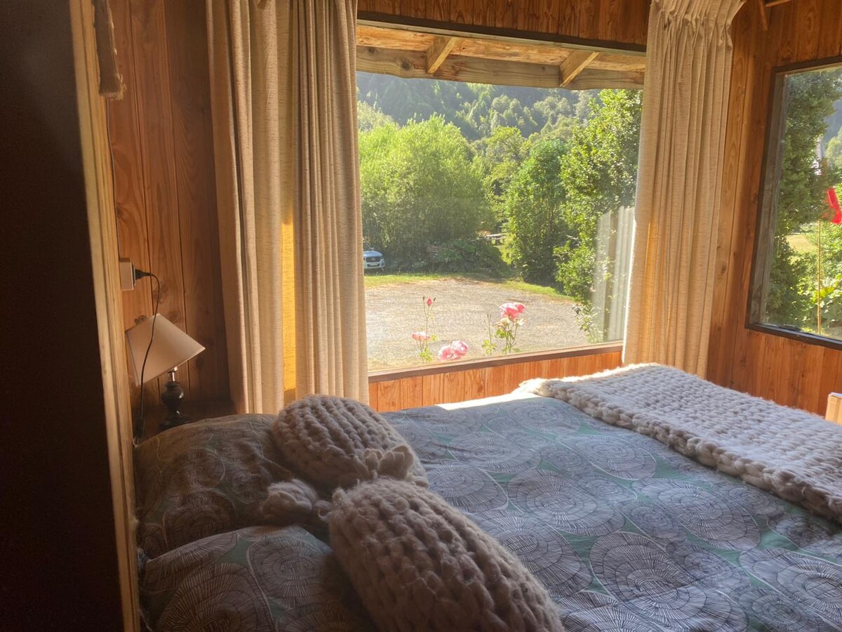 A cozy bedroom features a spacious bed adorned with a textured blanket. Natural light floods in through a large window, which frames a view of greenery outside. Soft curtains drape beside the window, and a bedside lamp adds a subtle touch of illumination.