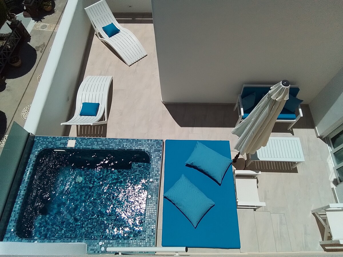 An aerial view showcases a private outdoor area featuring a sparkling jacuzzi surrounded by mosaic tiles. Two vibrant blue lounge cushions are positioned on a sunlit deck, accompanied by white loungers and a stylish umbrella, inviting relaxation in a tranquil setting.