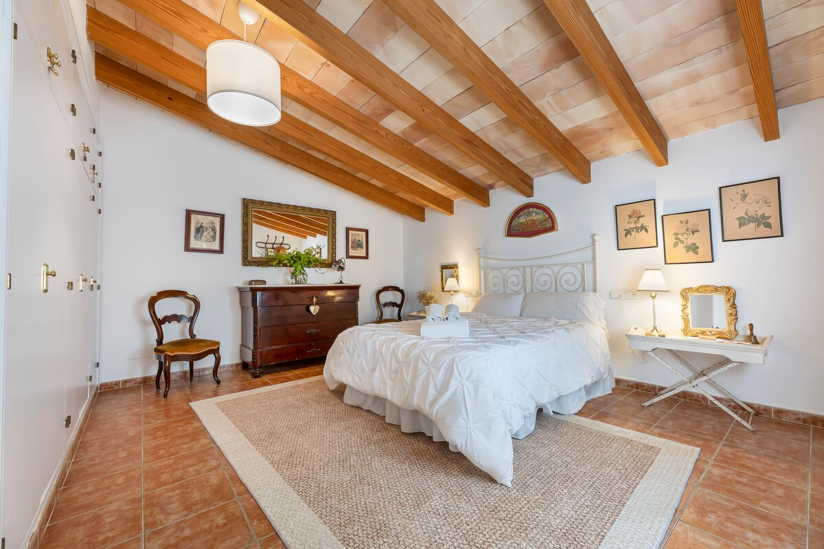 A spacious bedroom features a comfortable bed with a soft white coverlet. Wooden beams line the ceiling, complemented by vintage decor and softly lit lamps. A dresser and a small table provide additional furnishings, while botanical prints adorn the walls, enhancing the inviting feel of the room.