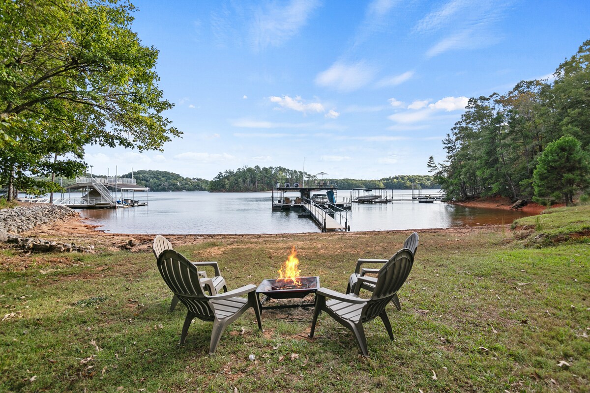 A serene outdoor space features two Adirondack chairs arranged around a fire pit, set against the backdrop of Lake Lanier. The calm water reflects the sky, while a dock and boats are seen in the distance, inviting leisurely moments by the lake.