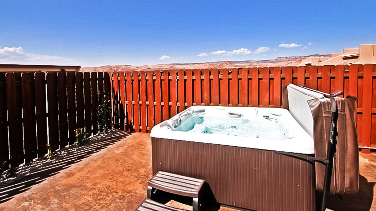 A private hot tub is positioned on a concrete patio, surrounded by wooden fencing. Clear blue skies and distant desert landscapes are visible, providing a serene setting for relaxation. A wooden bench sits nearby for convenience.