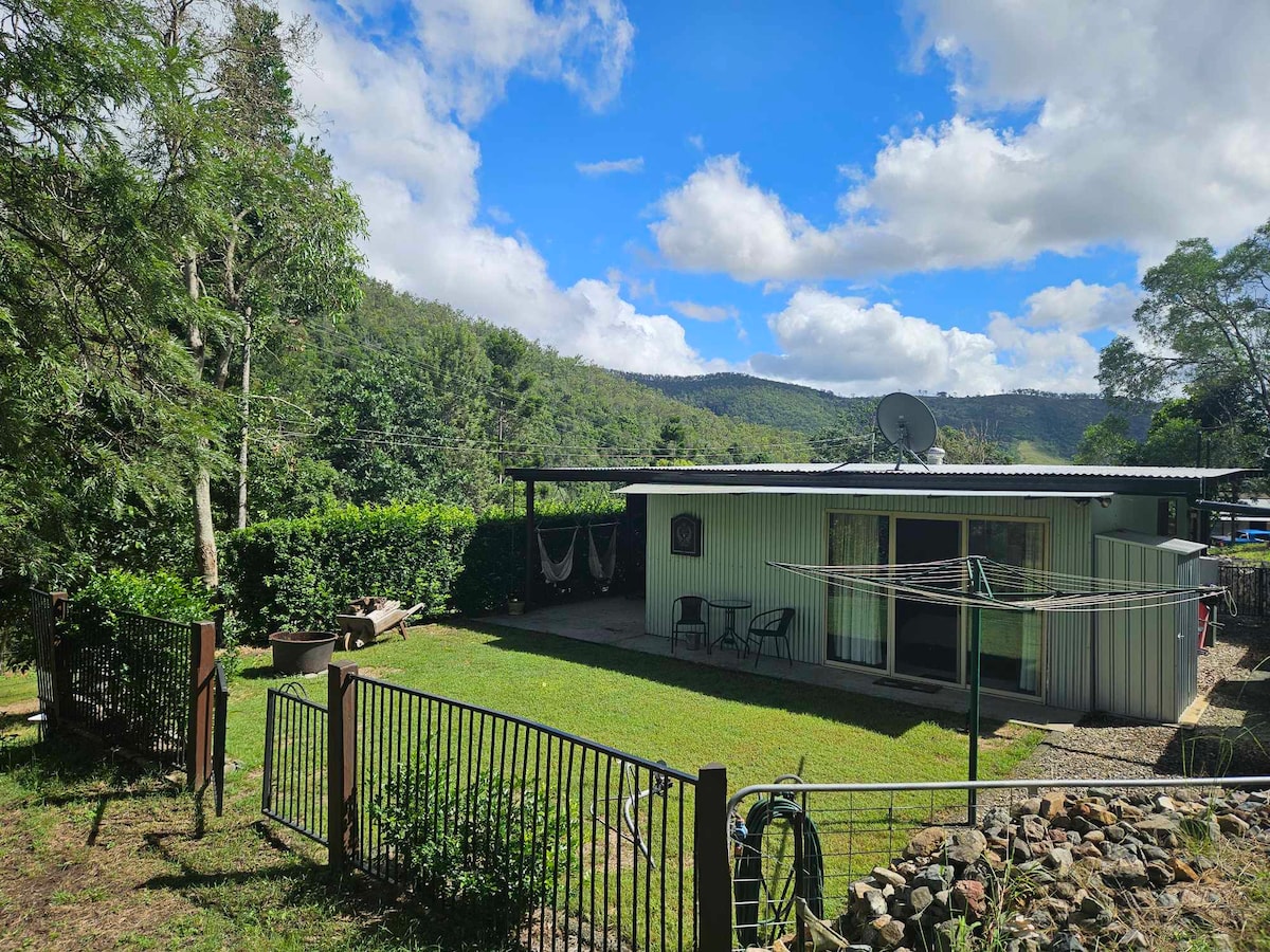 A quaint cabin is surrounded by lush greenery and rolling hills, featuring a well-manicured lawn. Outdoor seating is provided in front, with a clothesline and a fire pit visible. Bright blue skies and scattered clouds create a serene atmosphere in this natural setting.
