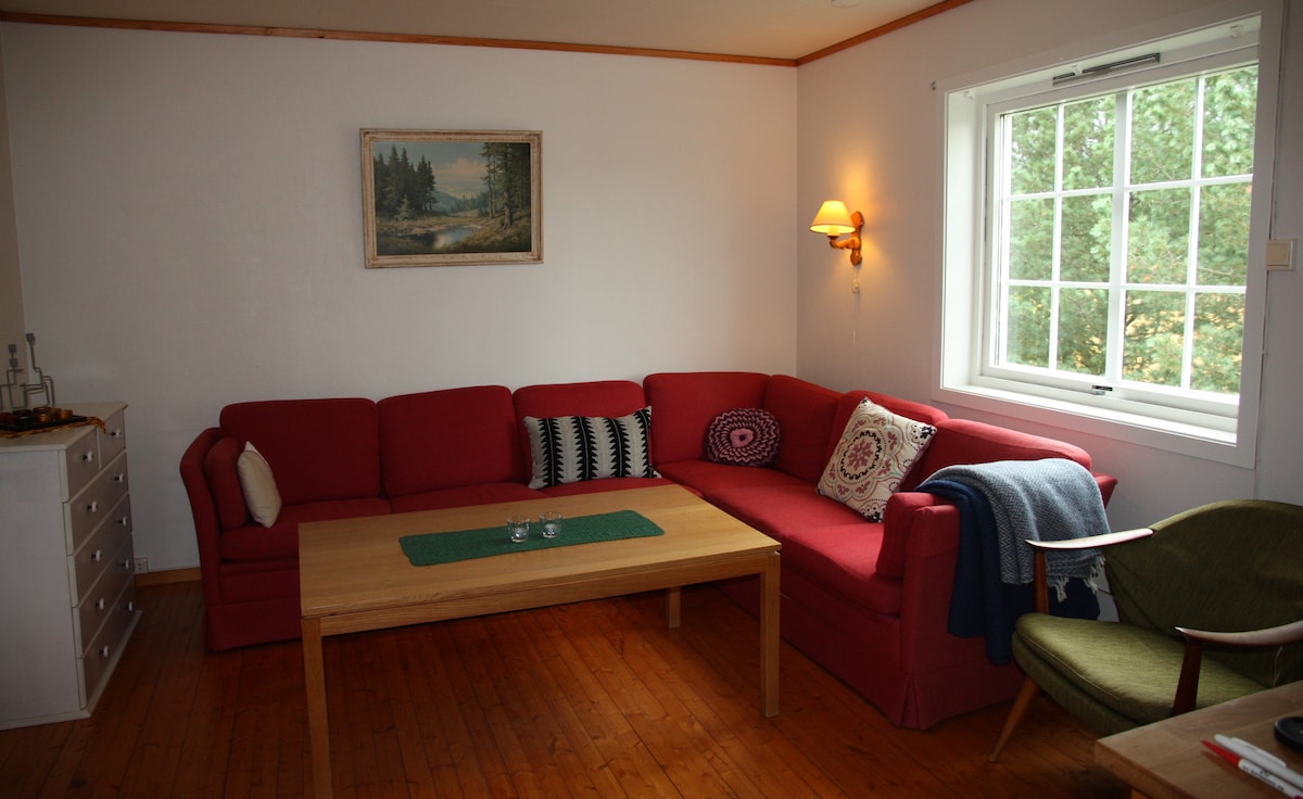 A comfortable living area features a red sectional sofa adorned with decorative cushions. A wooden coffee table sits centrally, complemented by a green table runner. Natural light filters through a large window, which offers views of the surrounding greenery. A cozy armchair and a painting enhance the ambiance.