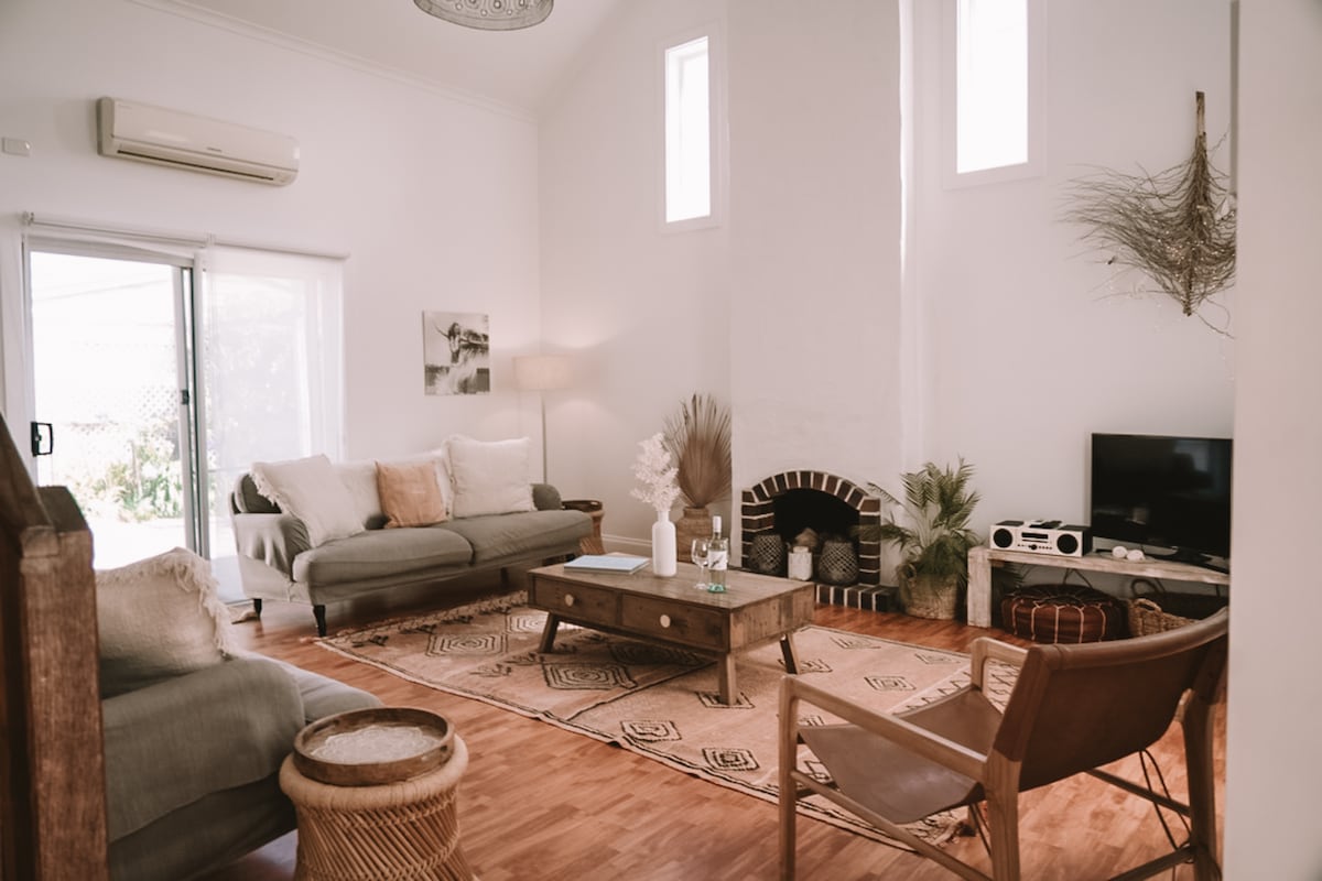 A bright living area features high ceilings and large windows, allowing natural light to fill the space. Comfortable seating is arranged around a wooden coffee table, with a rug placed on polished wooden floors. A fireplace adds a cozy touch, while decorative elements enhance the welcoming ambiance.