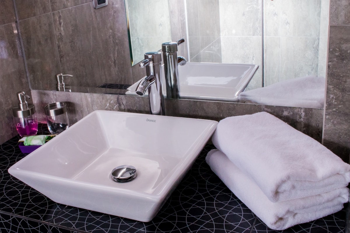 A modern bathroom features a sleek, rectangular sink positioned on a dark countertop adorned with a geometric pattern. Soft white towels are neatly stacked beside the sink. A large mirror reflects the stylish fixtures, creating a contemporary and clean appearance.