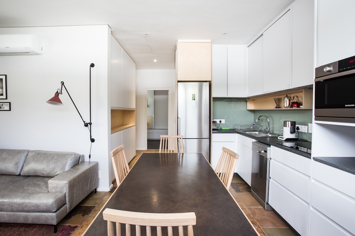A modern kitchen and dining area is visible, featuring a long table surrounded by wooden chairs. Light-colored cabinetry and a spacious countertop create a clean look. A comfortable sofa sits nearby, while appliances are neatly integrated, enhancing the functionality of the space.