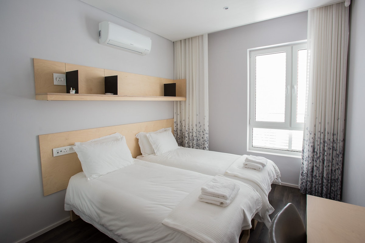 A well-lit bedroom features two spacious beds positioned side by side, each with white linens and a folded towel on top. A wooden shelf above the beds holds two black speakers and a decorative item. Natural light enters through a large window, enhancing the modern design.