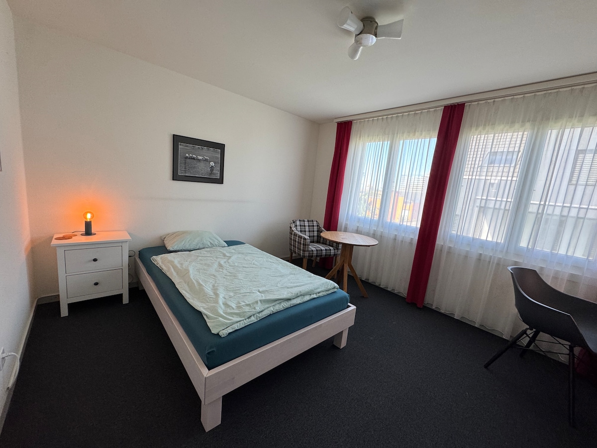 A modern guest room features a spacious bed with light bedding, complemented by a bedside table with a lamp. A checkered chair sits next to a round table, while a desk with a chair is positioned near large windows draped with sheer curtains.
