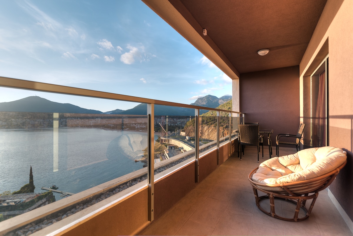 A spacious terrace offers panoramic views of the sea and nearby mountains. Comfortable seating arrangements, including two black chairs and a round, cushioned lounge chair, enhance the outdoor space. Large glass railings provide an unobstructed view of the surrounding landscape, inviting relaxation.