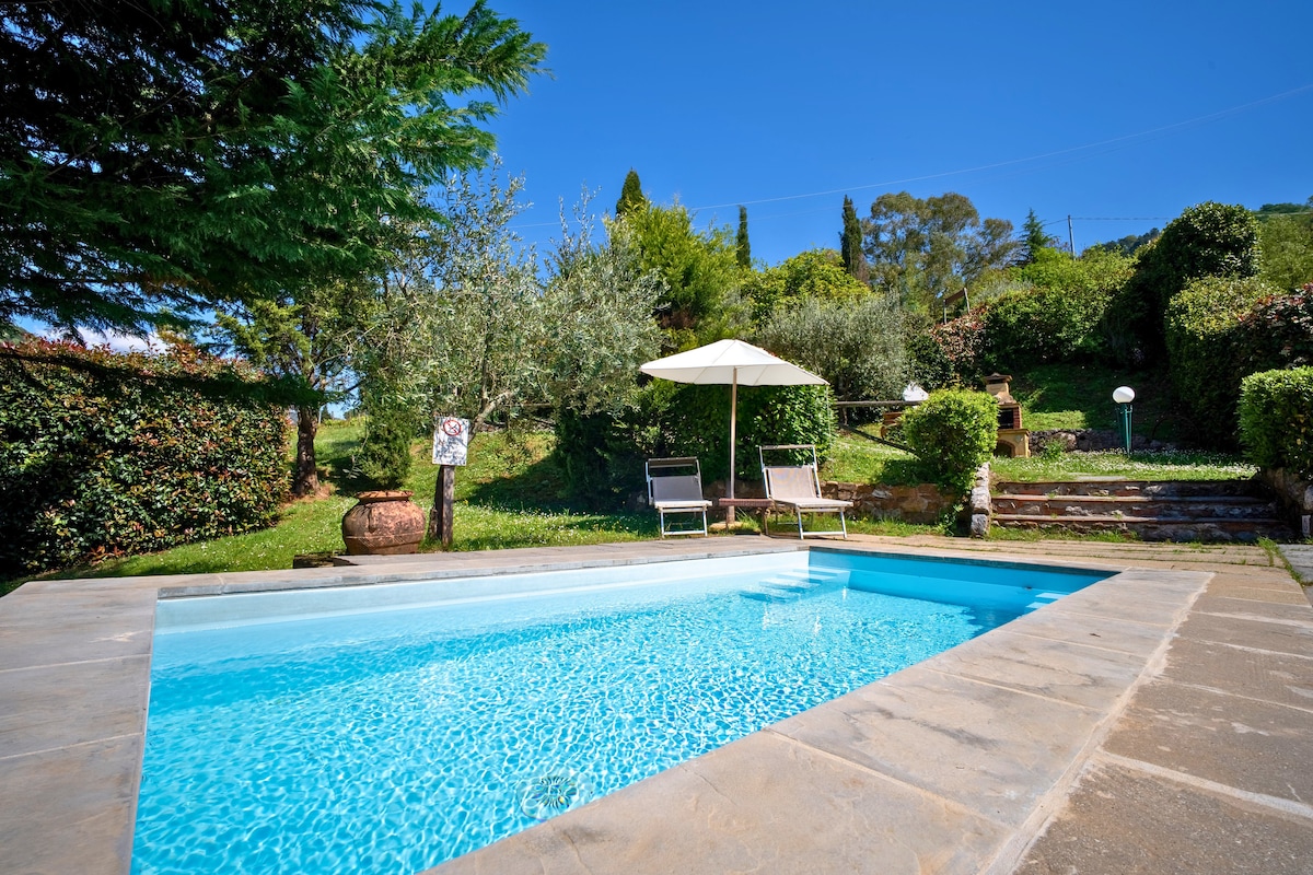 A private swimming pool is surrounded by lush greenery and soft grass. Sun loungers sit beside the pool, and a shaded umbrella stands nearby. Stone steps lead up to a well-maintained garden area, inviting relaxation in a serene environment.