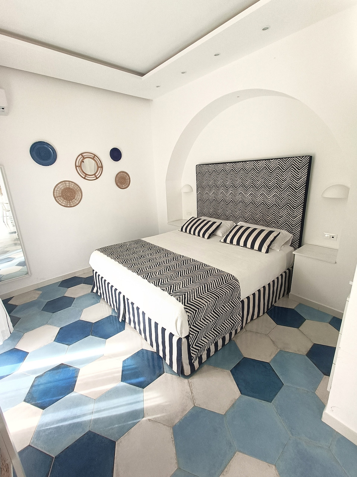 A spacious bedroom features a king size bed with a patterned headboard and coordinating striped bedding. Decorative wall baskets add a touch of texture. The hexagonal tile flooring in varying shades of blue and white enhances the serene ambiance. Natural light enters through the window.