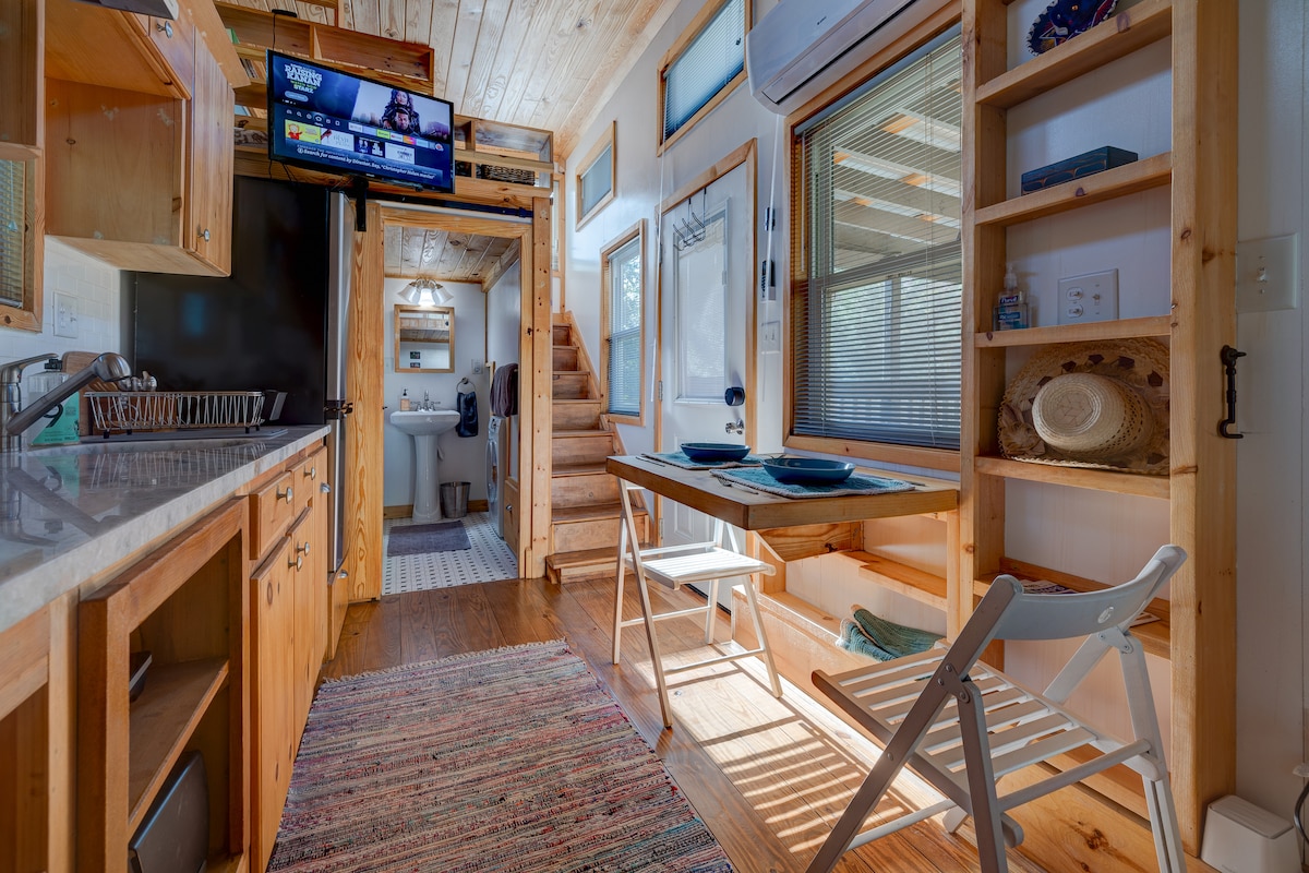 The interior of the tiny house is spaciously arranged, featuring a sleek kitchenette with wooden cabinetry and a small, pull-out dining table beside it. Natural light fills the area, highlighting the wooden walls and open layout, which includes a staircase leading to the sleeping loft.