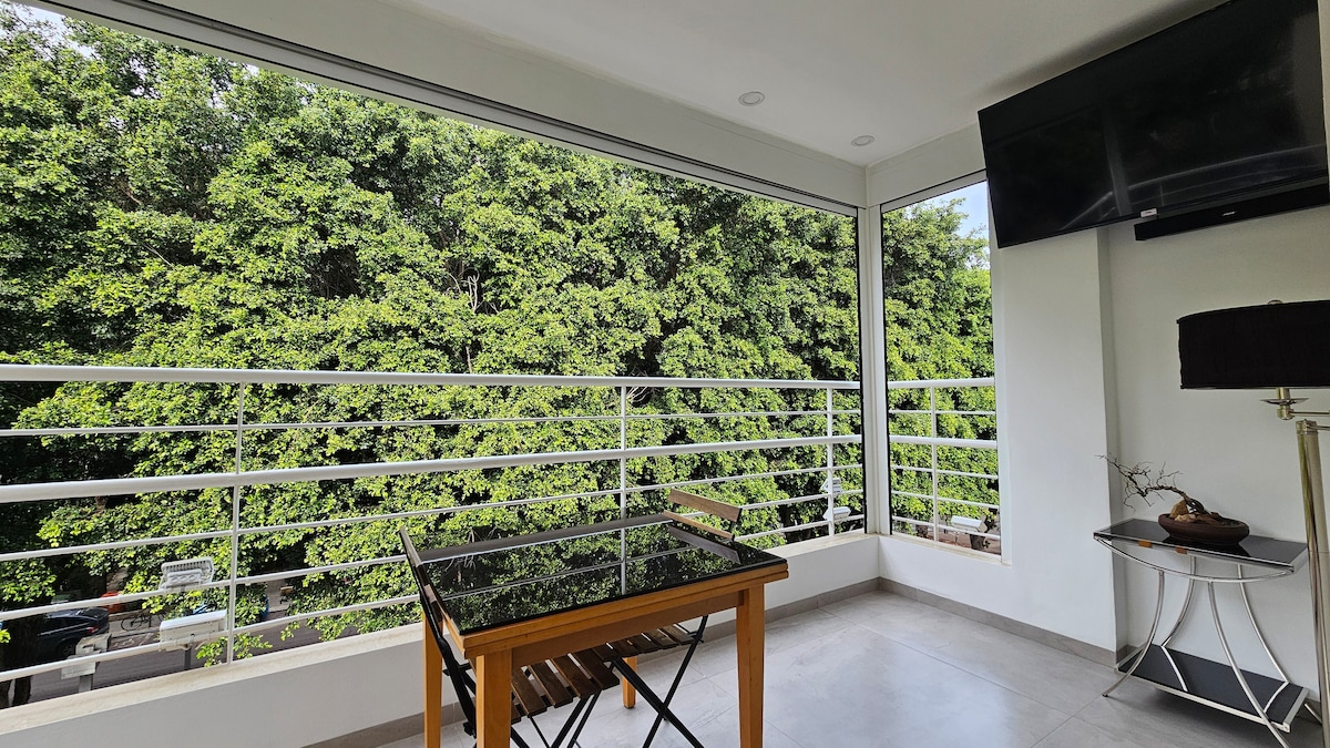 A balcony is showcased with a glass table and wooden chairs, overlooking a view of lush green foliage. Large windows allow ample natural light into the space, and a mounted television is positioned on the wall beside decorative lighting.
