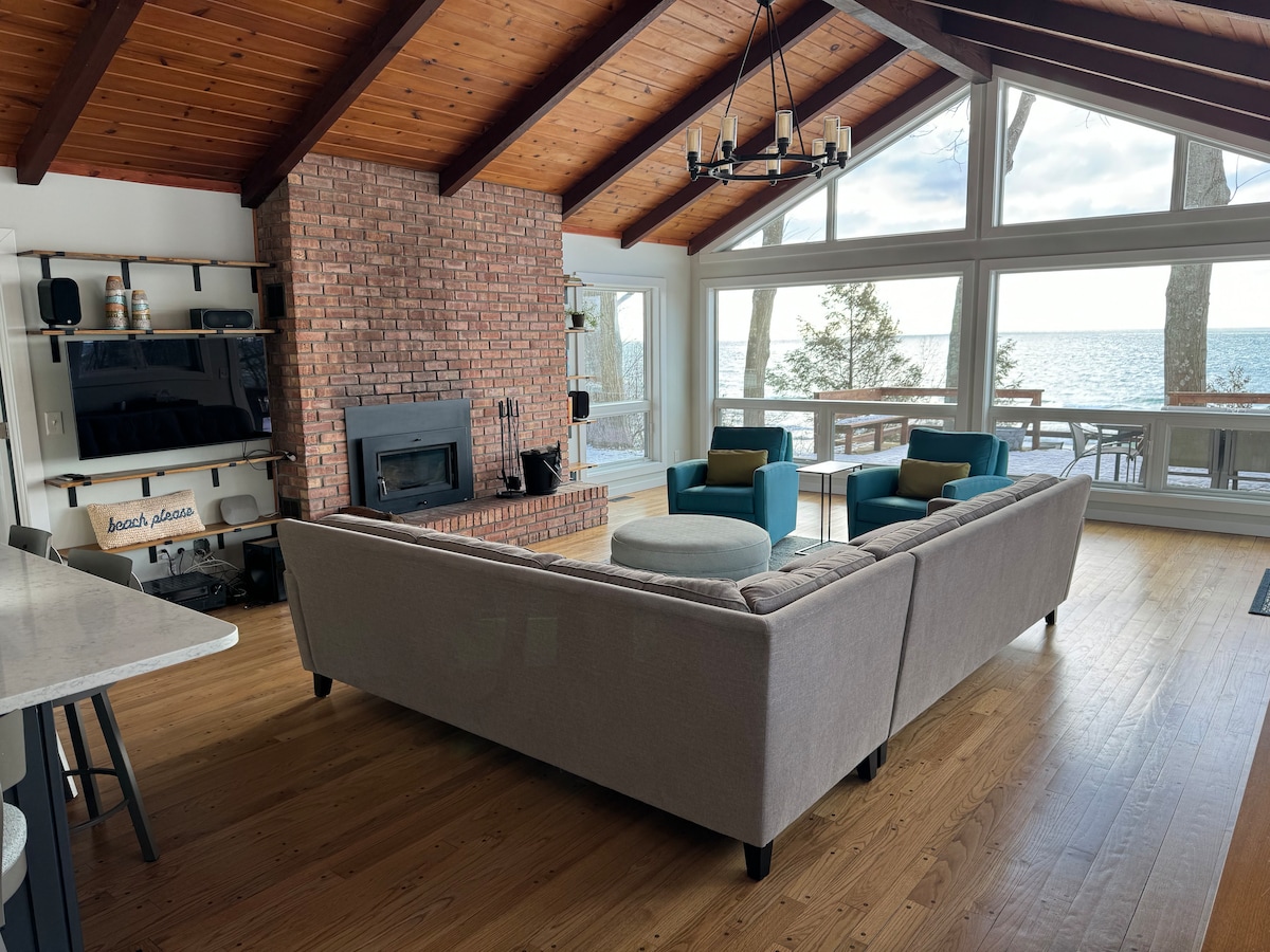 The spacious living area features a high, wood-paneled ceiling and large windows offering views of the lake. A comfortable sectional sofa is positioned around a rustic brick fireplace. Two accent chairs in a soothing hue complement the warm tones of the wood flooring.
