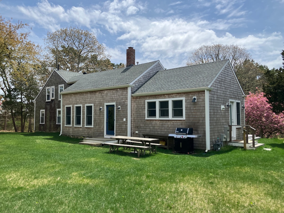 The exterior of the farmhouse features a shingled facade, complemented by large windows that allow natural light. A spacious yard, filled with green grass, includes a picnic table and a Weber grill on a wooden deck. Trees and blooming shrubs are visible in the background.