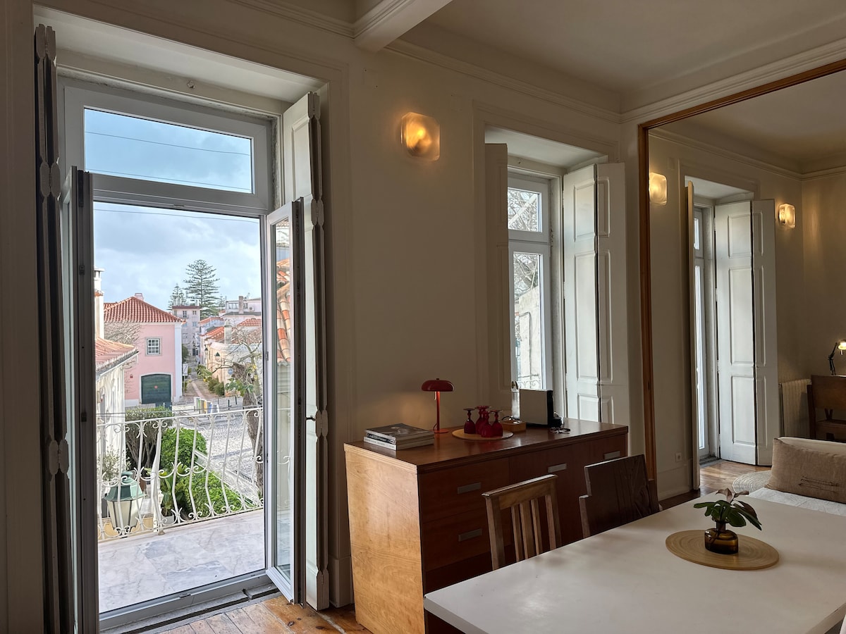 Natural light fills the open space, showcasing a dining area with a circular centerpiece and wooden chairs. Two large windows are open, allowing fresh air and offering views of the surrounding neighborhood. A wooden sideboard complements the modern decor.