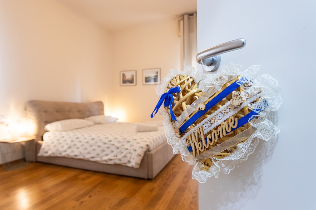 A heart-shaped welcome sign, adorned with blue ribbons and lace, hangs on the door. In the background, a spacious bed is visible, accompanied by soft lighting that creates a soothing ambiance.