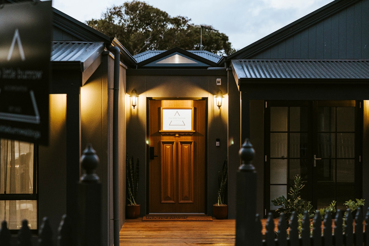The entrance to the little burrow is framed by warm exterior lighting, highlighting a charming wooden door adorned with a geometric design. Flanking the door are potted plants, and a welcoming deck leads to the inviting entryway.