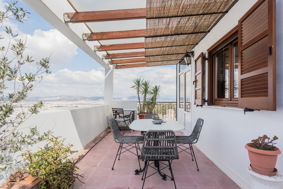 A spacious private terrace features a round dining table surrounded by six chairs, ideal for outdoor meals. Potted plants and greenery are present, enhancing the setting. Wooden beams and a retractable awning provide shade while showcasing sweeping views of the city skyline and hills.