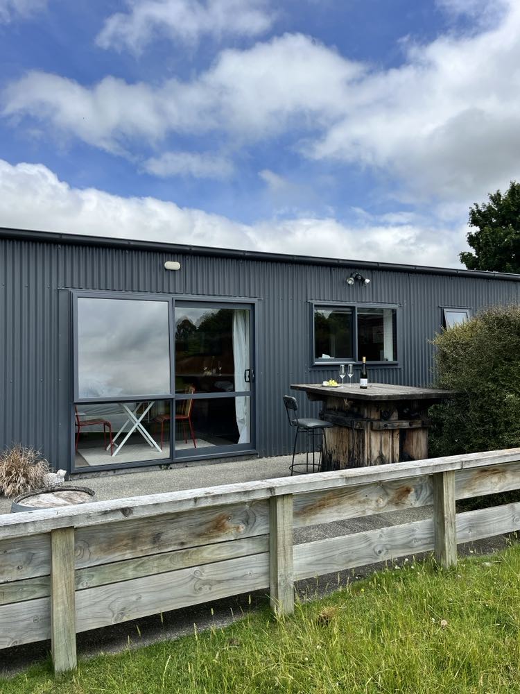 An outdoor view of the self-contained unit reveals a sleek, modern facade with large glass doors. A small patio area features a rustic table and chairs, surrounded by greenery and a wooden railing, inviting relaxation in the fresh air.