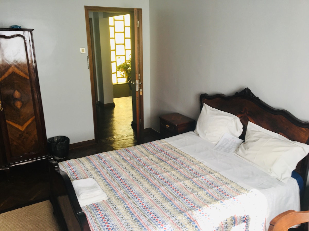 A spacious bedroom features a wooden bed frame with a neatly made bed, complemented by decorative pillows. A wardrobe is positioned against one wall, with a wastebasket nearby. A doorway is visible, leading to a well-lit area beyond.