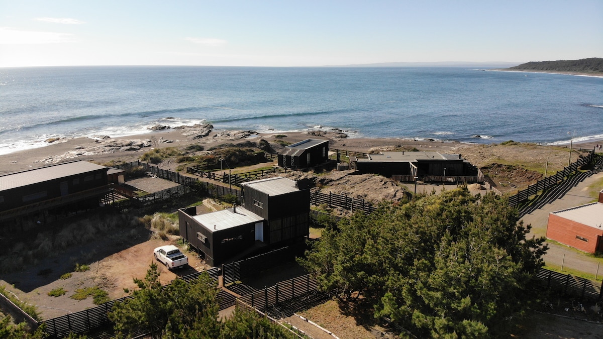 A panoramic view captures a collection of modern houses nestled along the coastline, with the ocean visible in the background. A sandy beach extends into the distance, while lush greenery surrounds the properties. The tranquil waters reflect the clear sky, creating a serene atmosphere.