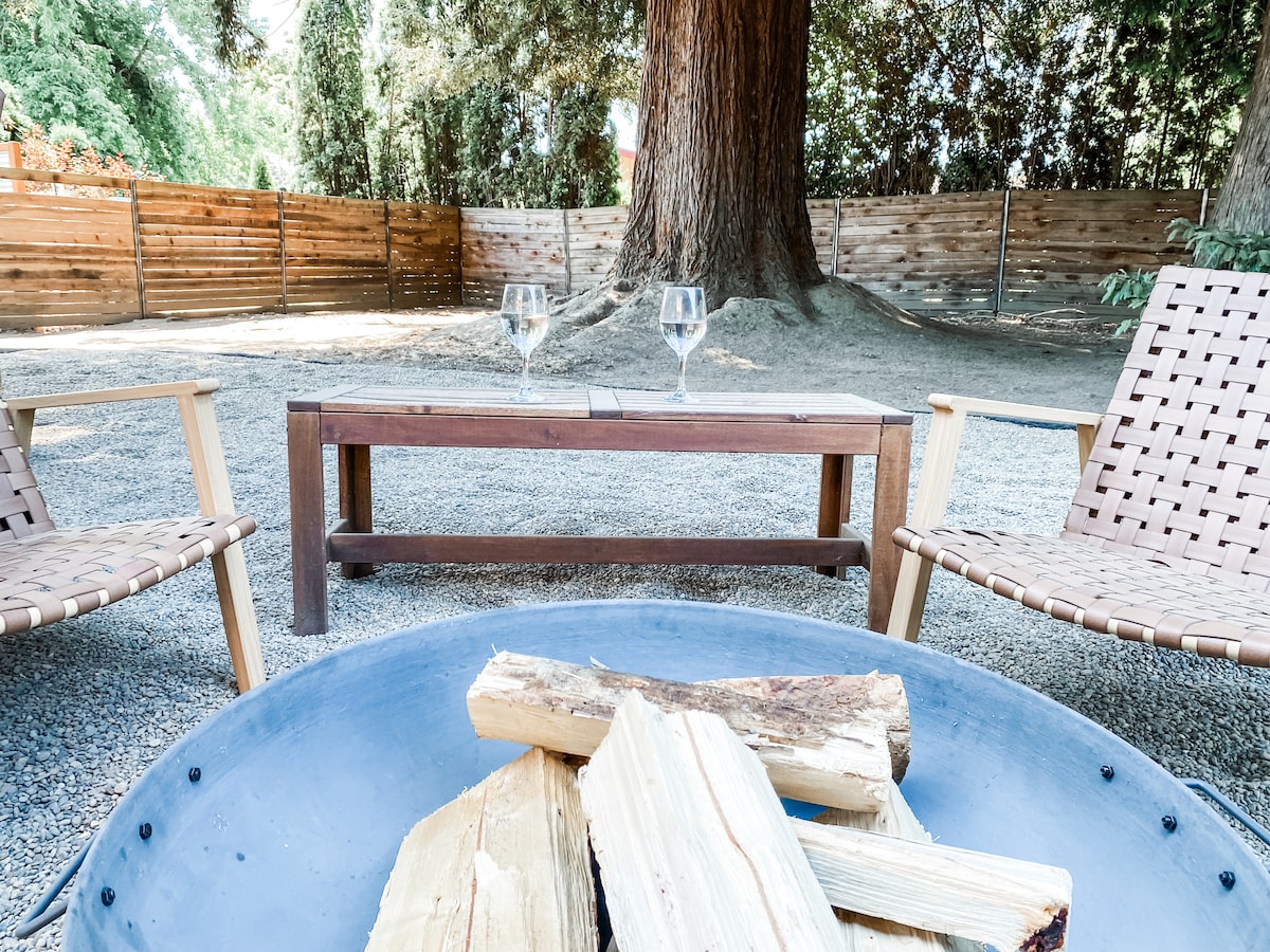 A cozy outdoor area is featured with two woven chairs positioned around a metal fire pit filled with logs. A wooden table sits between the chairs, set for two with wine glasses. A majestic Redwood tree provides shade in the background.