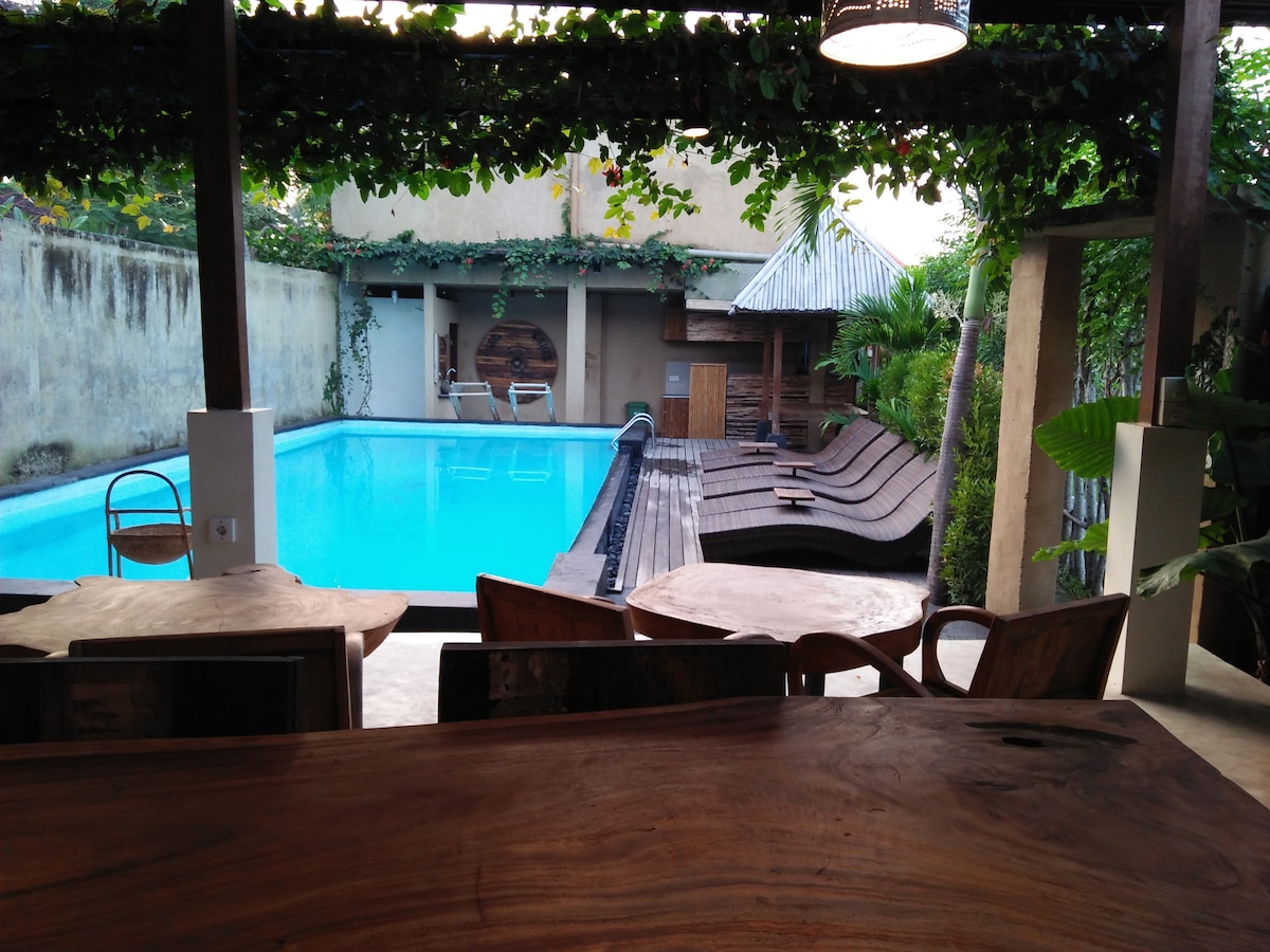 A serene view of the inviting pool area is captured, featuring soft lounge chairs arranged along the deck. The pool is surrounded by lush greenery, with a traditional straw-roof structure visible in the background, adding to the tranquil atmosphere.