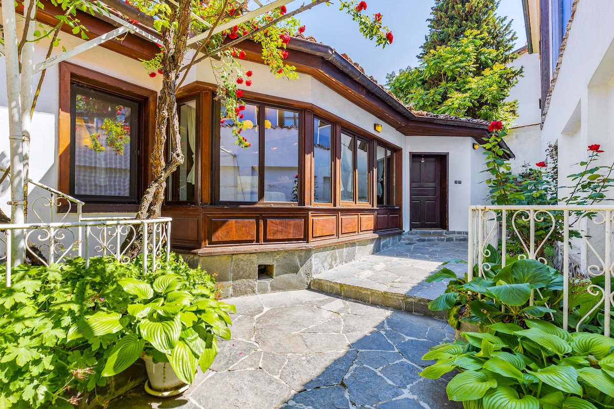 A stone pathway leads towards the house, surrounded by greenery and vibrant flowers. Large windows are framed with wooden accents, enhancing the entrance's inviting character. A wrought-iron railing enhances the charm, while trees provide a natural canopy overhead.