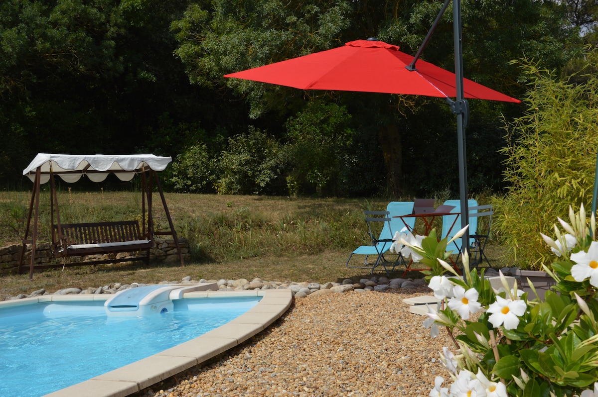 The image features a clear swimming pool accompanied by a red umbrella providing shade. A wooden swing and a small seating area with blue chairs are positioned nearby, surrounded by lush greenery and blooming flowers, creating a serene outdoor space.