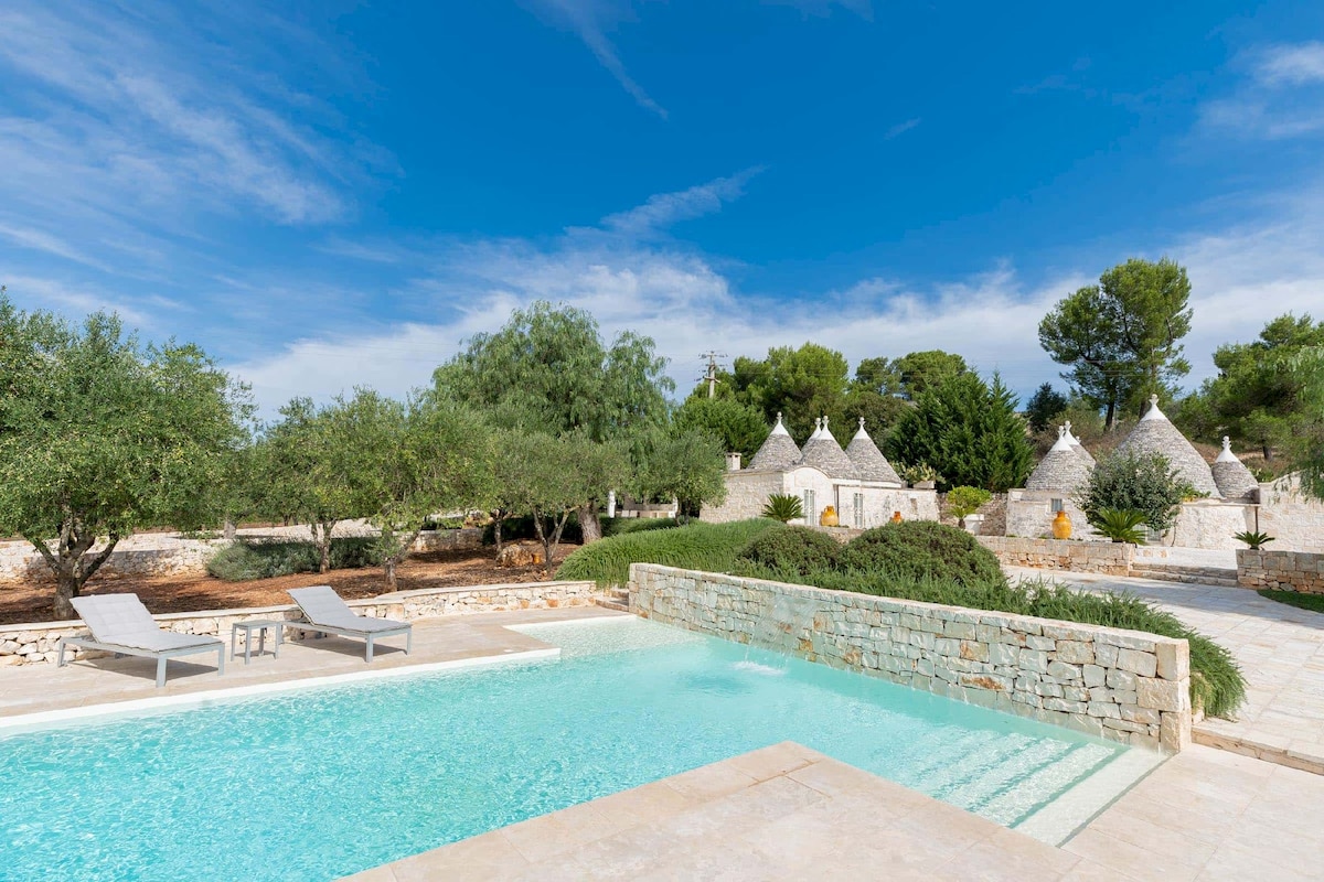 A serene saltwater infinity pool is surrounded by lush greenery and ancient trees. Comfortable lounge chairs are positioned nearby. Traditional Trullo houses are visible in the background, set against a backdrop of clear blue skies, adding to the tranquil countryside ambiance.