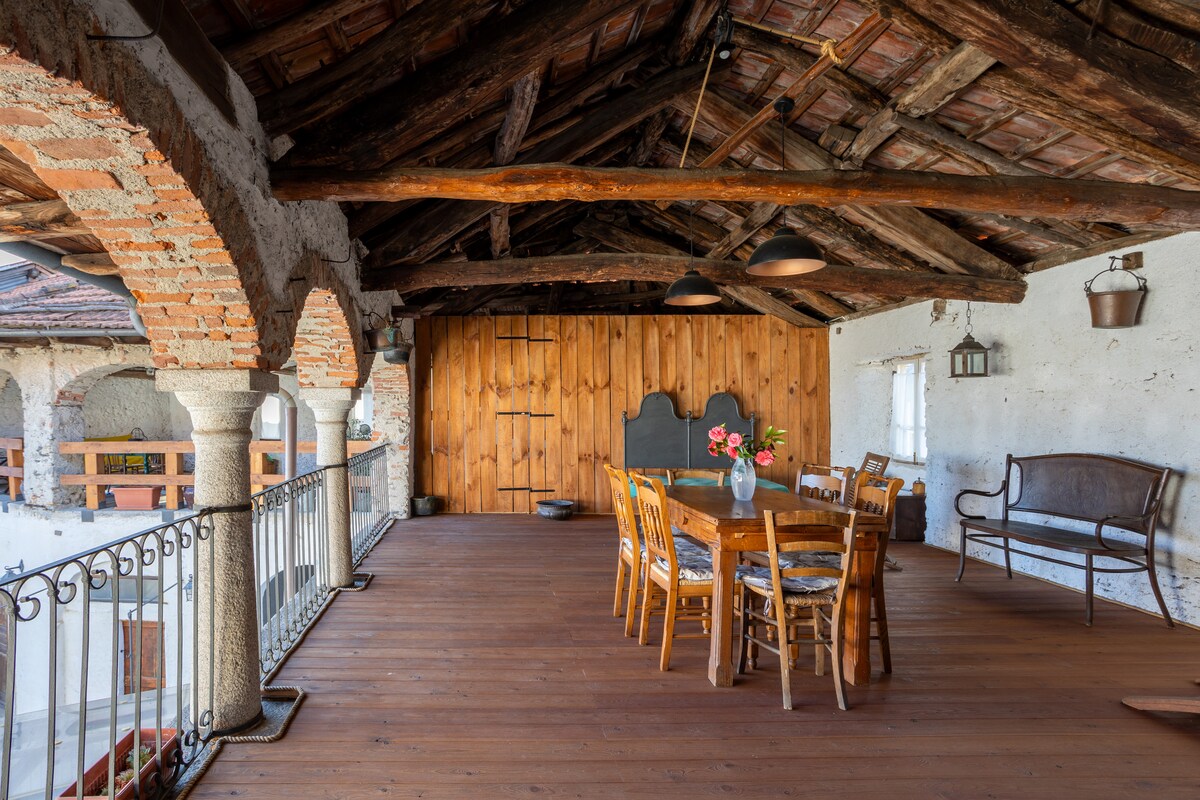 An outdoor loggia is depicted, featuring a large wooden dining table surrounded by chairs. The rustic wooden beams and terracotta tile roof enhance the authentic character, while a small vase with flowers adds a touch of vibrancy. A bench lines one side, and a railing provides a view.