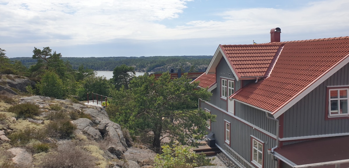 A charming gray house with a red roof stands on rocky terrain, surrounded by trees. Views of the water and distant forest are visible in the background, highlighting the serene landscape of Sannäs.