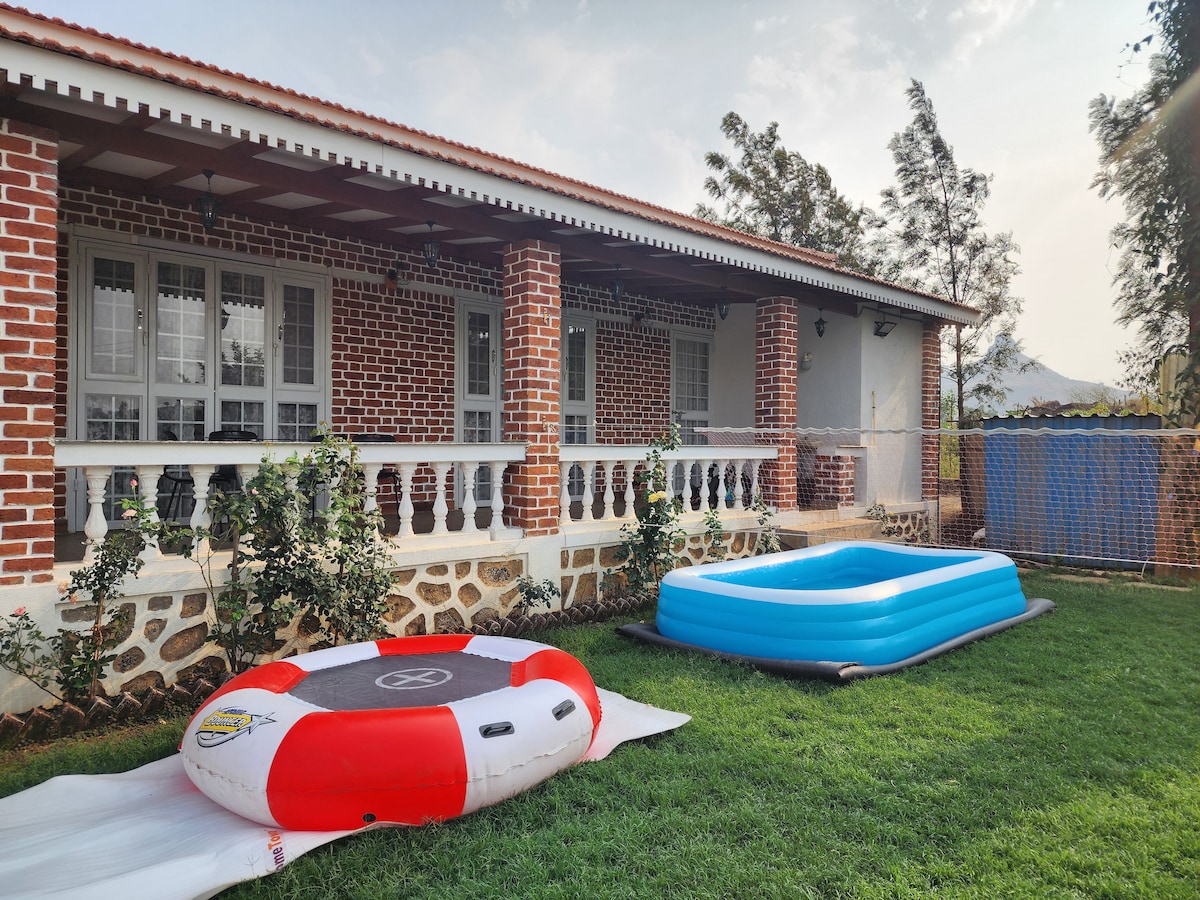 The outdoor area features two inflatable pools, one circular with red and white accents and another rectangular in blue. A spacious lawn surrounds the pools, and a charming brick facade of the villa is visible, complemented by large windows and a porch with decorative columns.