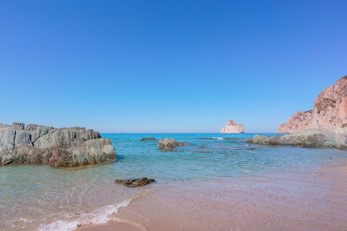A serene beach scene is captured, featuring gentle waves lapping at the shore. Smooth rocky formations protrude from the clear blue water, while a distant coastal rock formation rises against the bright blue sky.