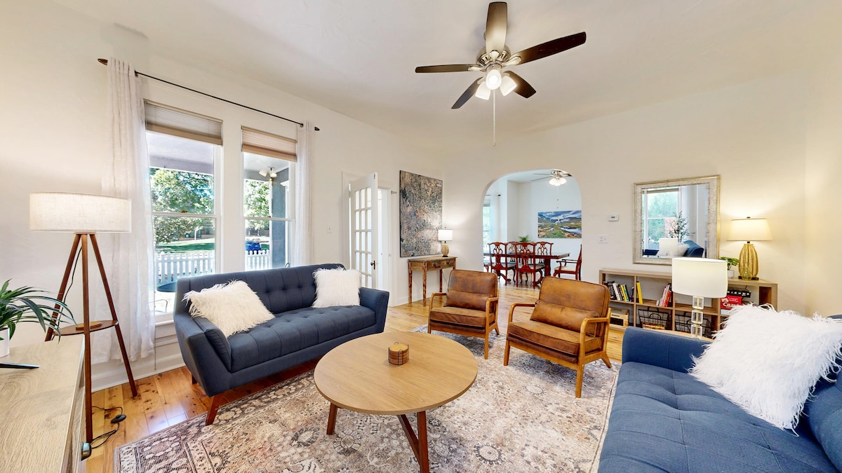 A spacious living room features two comfortable sofas and vintage armchairs arranged around a central coffee table. A ceiling fan and natural light from large windows enhance the inviting space. A dining area is visible in the background, showcasing a wooden table surrounded by chairs.