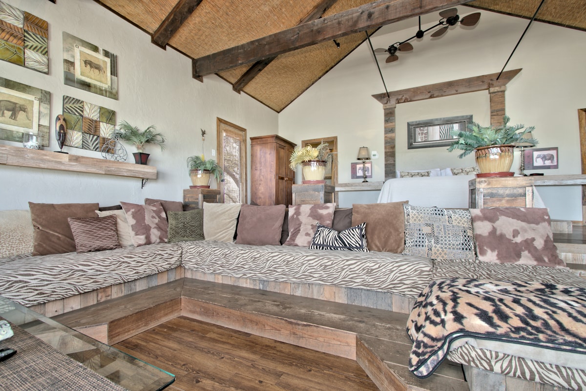 A spacious living area features a large, inviting sectional sofa adorned with a mix of animal prints and neutral-toned cushions. Natural light is welcomed through large windows, enhancing the rustic wooden accents and decorative shelves with plants and framed artwork.