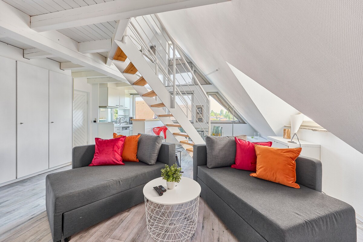 A modern living area features two gray couches adorned with colorful cushions in orange and pink. A round coffee table sits at the center, while large windows allow natural light to fill the space. A wooden staircase leads to the upper level.