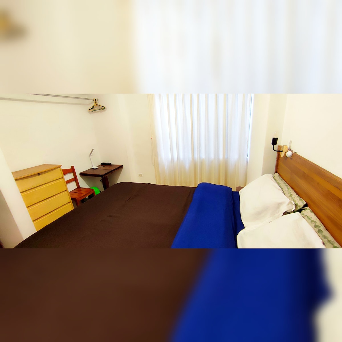 The room features a spacious bed with a dark coverlet and a blue throw blanket. A wooden dresser is positioned next to the bed, along with a chair and a small work desk. Natural light filters through sheer curtains, creating a bright ambiance.