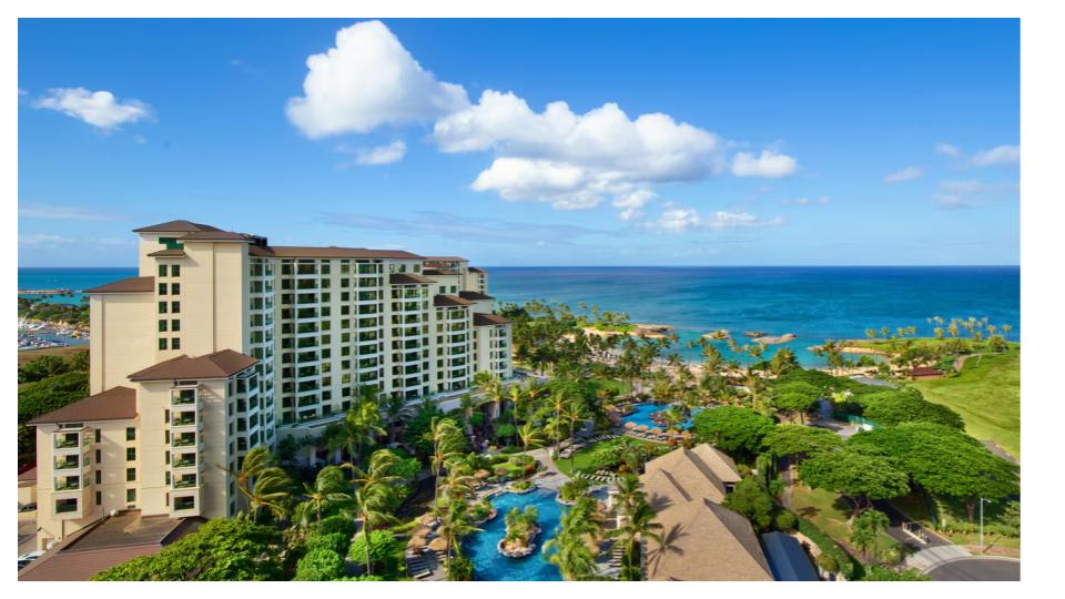 The image depicts a spacious resort complex nestled among lush greenery, with multiple levels and a sandy beach visible nearby. A sweeping ocean view reveals gentle waves under a partly cloudy sky, while the surrounding landscape includes palm trees and inviting swimming pools.