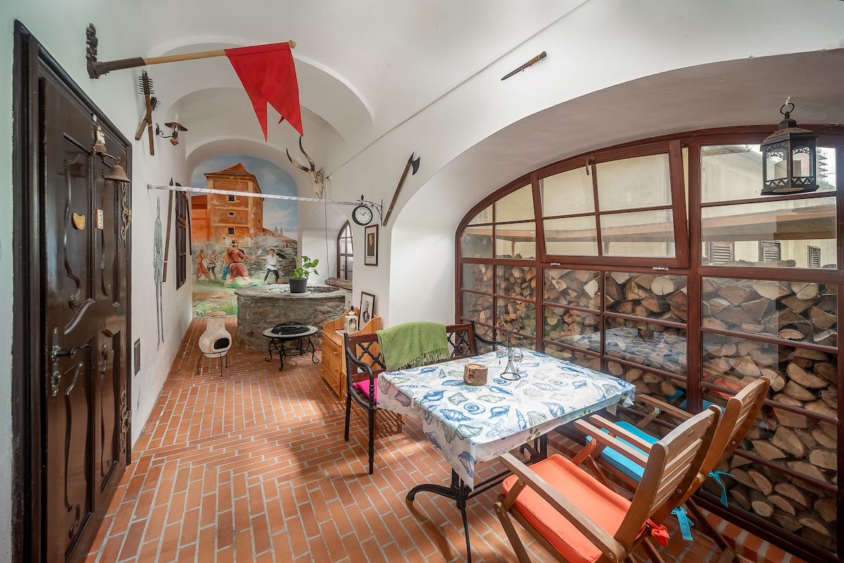 A cozy indoor dining area is presented, featuring a rustic wooden table surrounded by several chairs. The brick flooring complements the warm ambiance, while a mural depicting a historical building adds character to the space. A wall filled with stacked firewood enhances the inviting atmosphere.