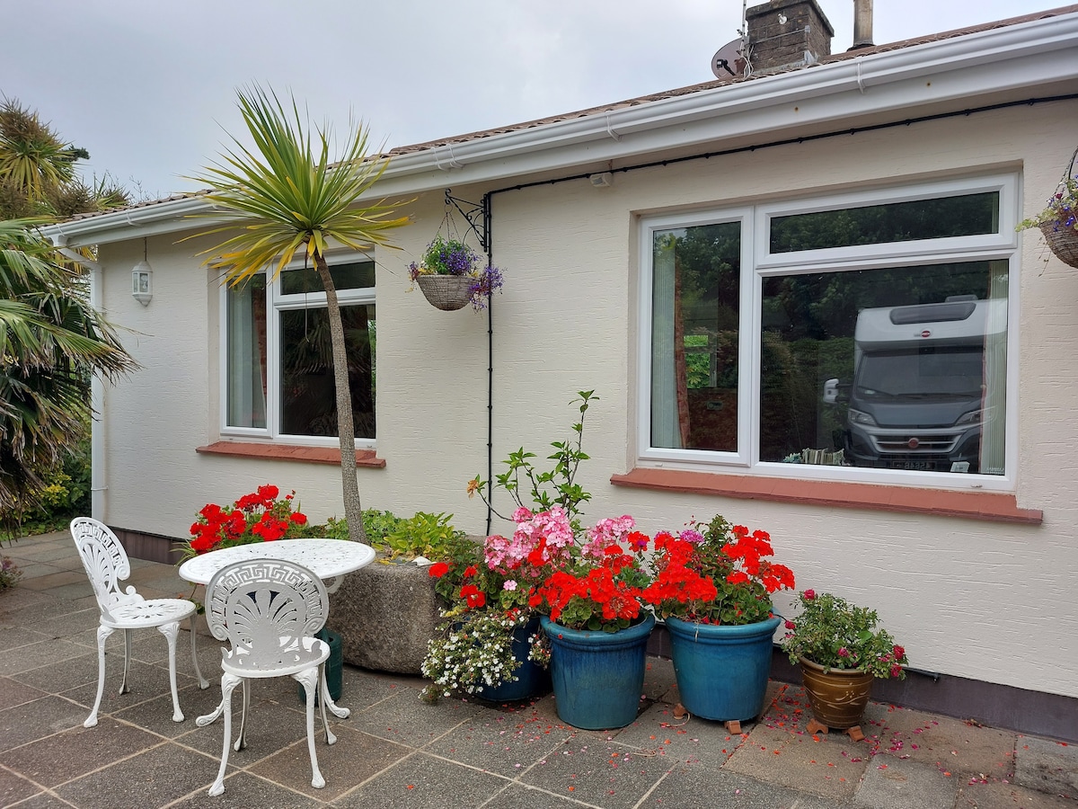 A charming exterior view of a bungalow showcases two large windows framed by colorful flower pots. A small round table with two white chairs is positioned on a stone patio, surrounded by vibrant red and pink blossoms, enhancing the inviting outdoor space.