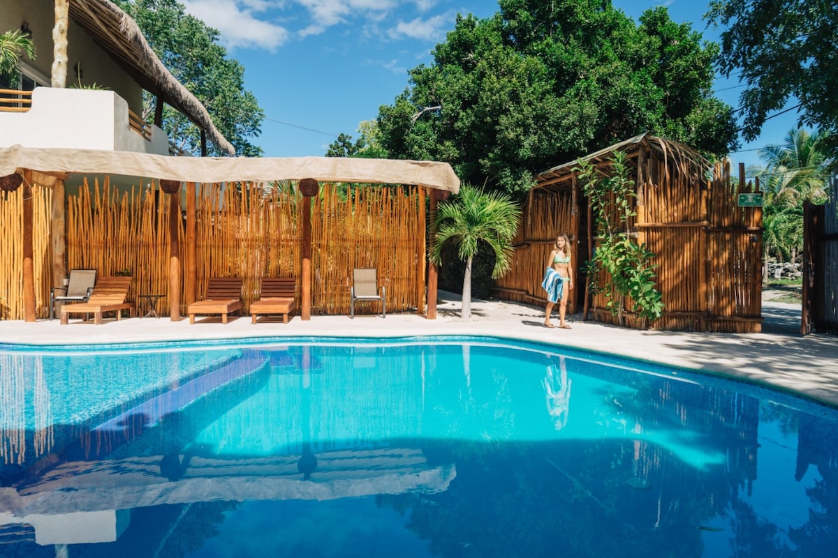 A well-maintained swimming pool is surrounded by a natural bamboo fence, with lounge chairs positioned nearby under sunshade. Lush greenery is visible in the background, providing a serene outdoor environment for relaxation.