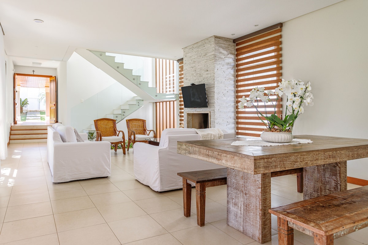 The open living and dining area showcases a wooden table surrounded by bench seating, complemented by white upholstered sofas. A flat-screen television is mounted on a stone wall, while large windows with wooden blinds provide natural light, creating a spacious and airy environment.