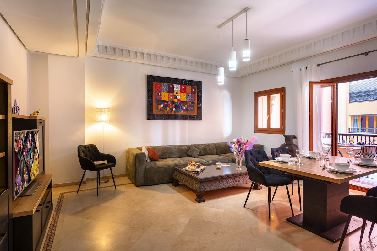 The living area features a comfortable sofa and a dining table set for four. Bright lighting is provided by pendant lights above, and a decorative artwork adorns the wall. Large windows allow for natural light and provide views of the outdoor terrace.
