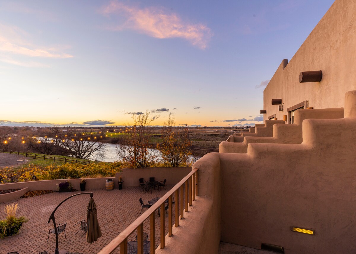 A spacious balcony offers views of the Yakima River and the sunset sky, featuring soft colors and distant clouds. The adobe-style exterior is complemented by a stone patio, which includes seating arrangements and landscaping with trees and shrubs.