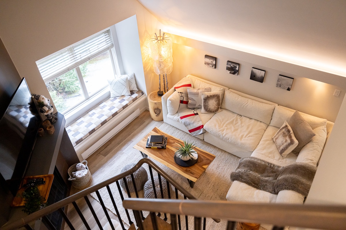 An inviting living area features a sectional sofa adorned with soft throw pillows. A wooden coffee table is placed at the center, complemented by a small plant. Natural light floods the space through large windows, and a cozy nook with a cushion offers additional seating.