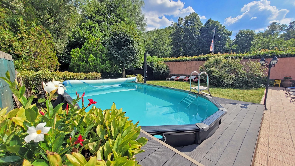 A serene swimming pool is surrounded by lush greenery, with comfortable lounge chairs positioned nearby. The pool area is complemented by flowering plants, and a gentle sunlight filters through the trees, creating a peaceful leisure space.