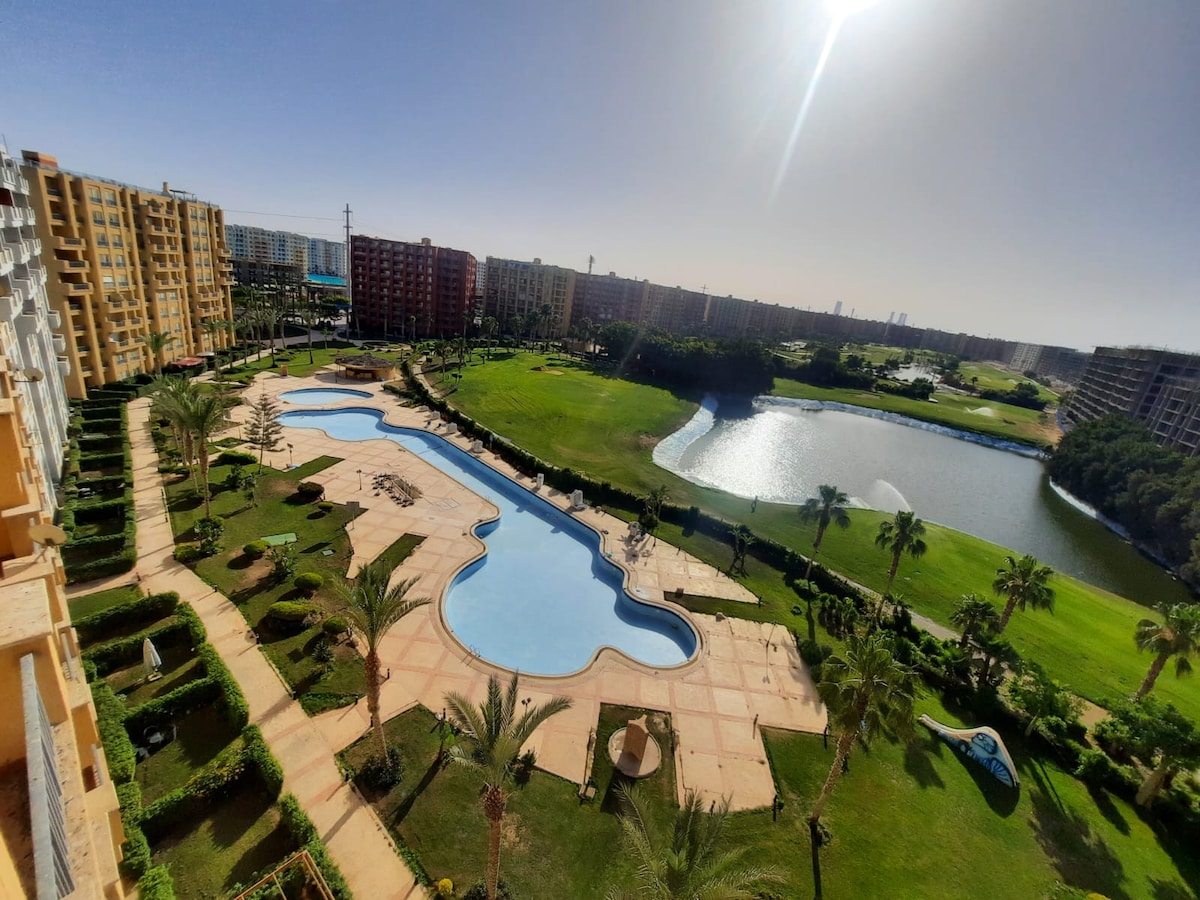 A large swimming pool is positioned amidst landscaped gardens, with palm trees lining the edges. Sun loungers and umbrellas are arranged on the poolside. A serene lake is visible in the distance, reflecting the clear blue sky, while paths meander through the lush greenery.