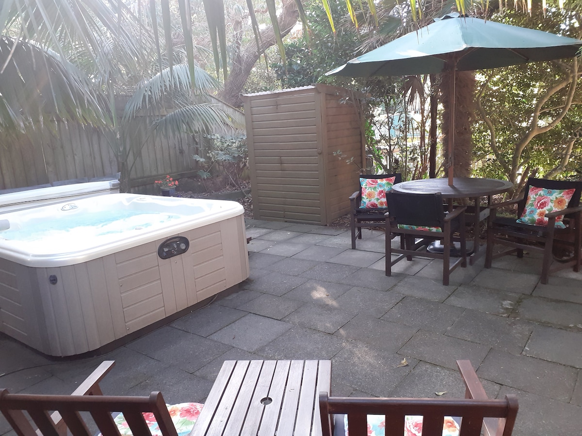 A private outdoor spa area is set among mature gardens, featuring a hot tub and a shaded patio table surrounded by four comfortable chairs. The space is paved, allowing for easy movement, with colorful cushions adding a touch of comfort.